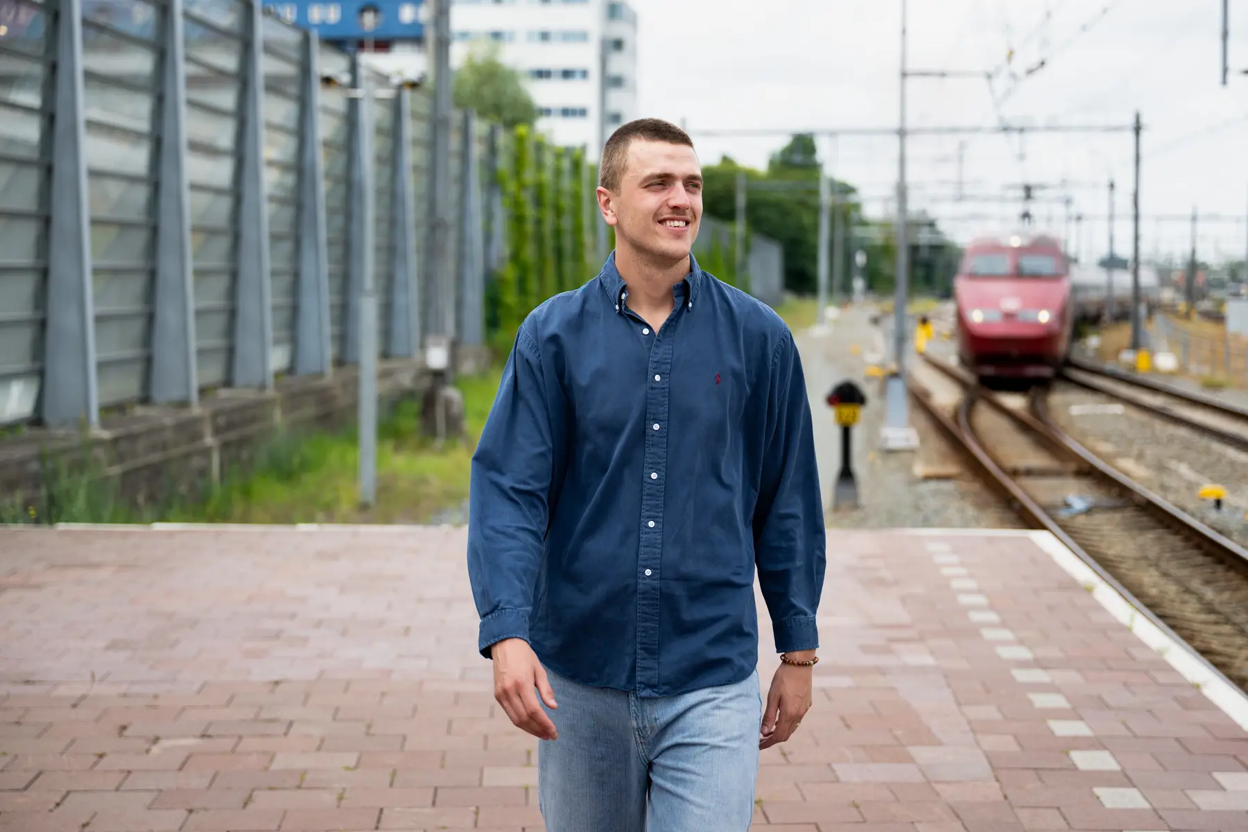 Bastiaan loopt op het perron naast het spoor, waar een trein op rijdt. Hij lacht en loopt er vrolijk bij, terwijl hij niet in de camera kijkt.