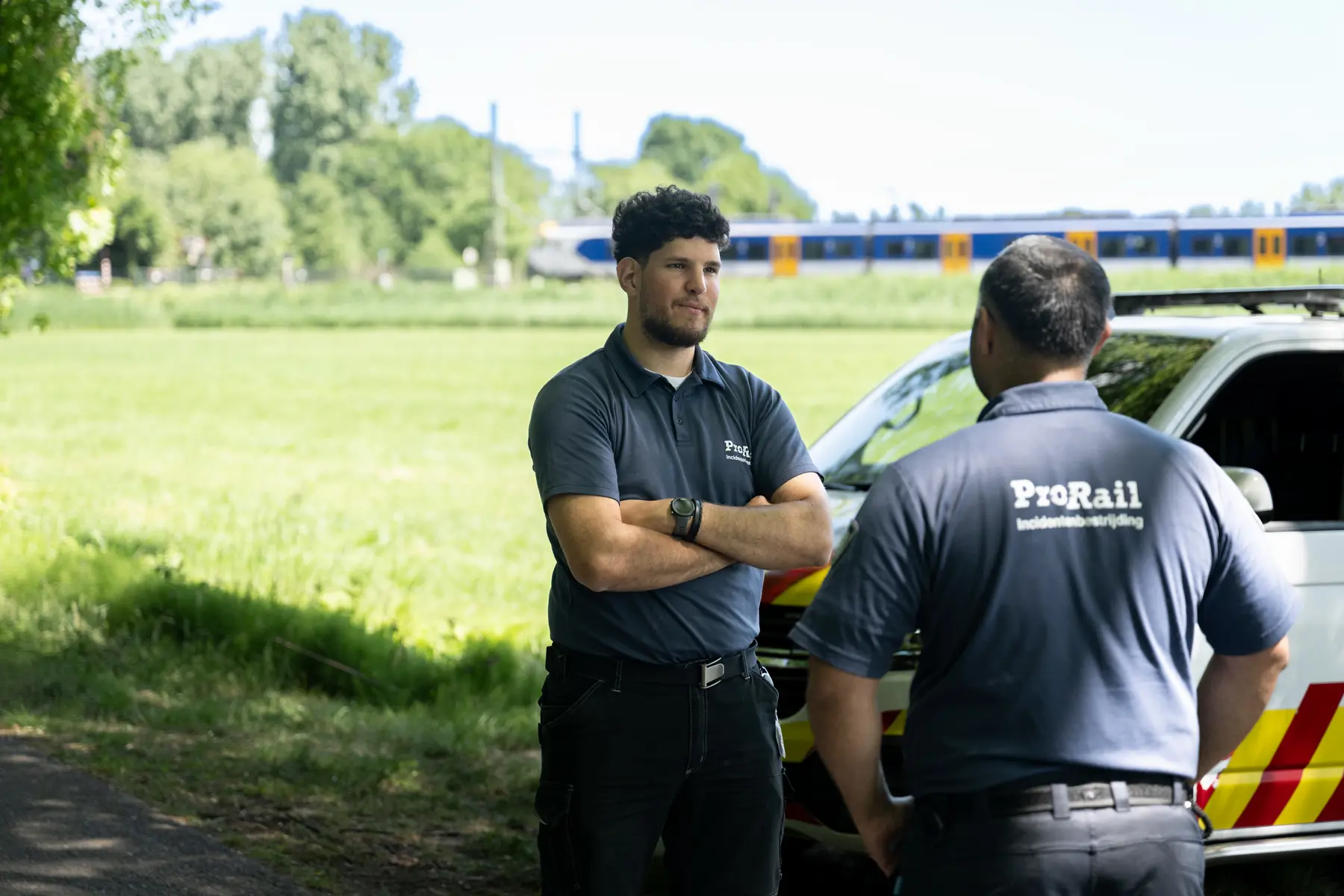 Stagiair Lucas Warmenhoven staat met een collega te praten, beide zijn van team Incidentenbestrijding. Ze staan naast een Incidentenbestrijdingbusje en op de achtergrond rijdt een trein.