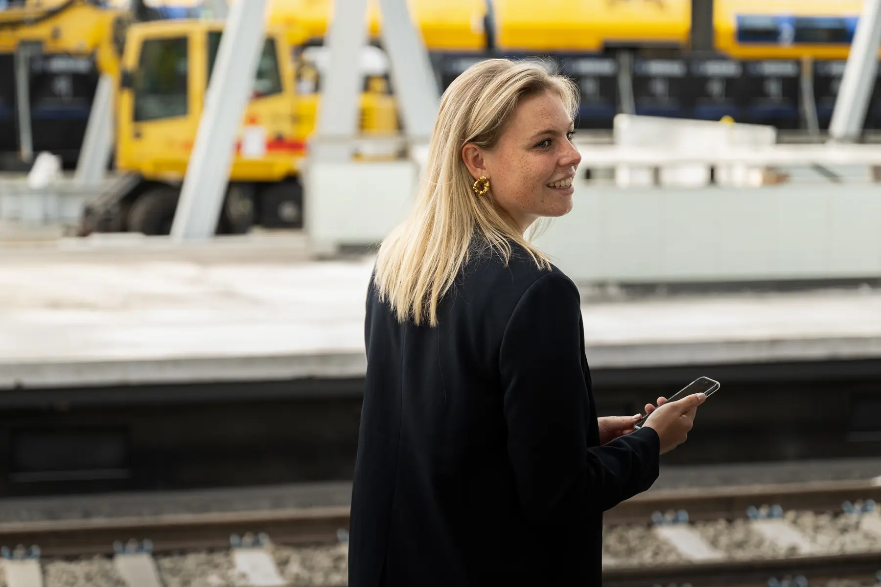 Merel Winters, technisch trainee, staat naast het spoor met de rug naar de camera. Ze kijkt over haar schouder en lacht. Ze heeft haar mobiel in de hand.